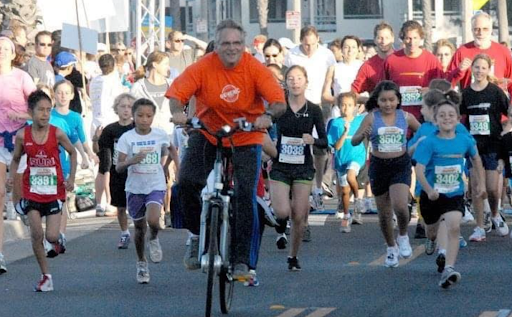Laurence Cohen cycling alongside young runners at a race